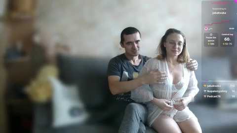 A video shows a man with short dark hair and a woman with light brown hair, both in casual attire, sitting closely on a couch. A blurred figure stands in the background.