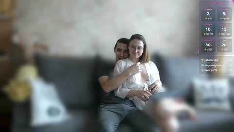 Video of a couple cuddling on a dark couch; woman with glasses, white top, jeans; man in black shirt, blue jeans. Background blurred.