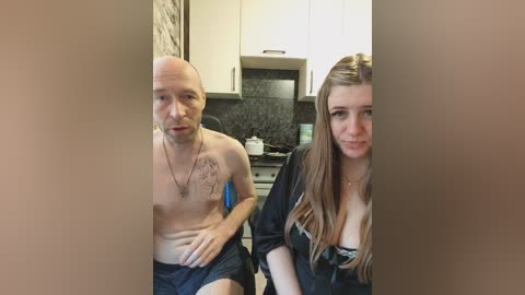 A video of a bald, shirtless man with tattoos, wearing shorts, sitting next to a woman with long brown hair in a black top, in a modern kitchen with white cabinets.