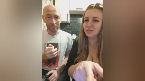 Video of a bald Caucasian man in a white T-shirt and a brunette Caucasian woman in a black top, both smiling in a modern kitchen with white cabinets and dark countertops.