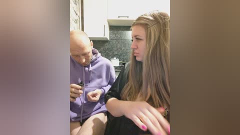 A video of a bald man in a purple hoodie and a young woman with long brown hair, seated in a modern kitchen with white cabinets and a granite countertop.