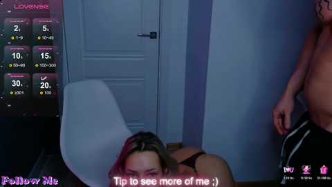 Video of a woman in black lingerie, face down on a bed, with a shirtless man standing nearby. The room is dimly lit, with a closed door in the background.