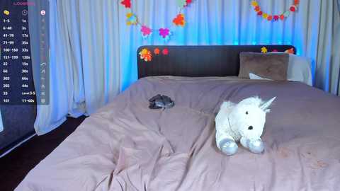Video of a bedroom with a plush unicorn toy on a bed, a cat resting on the bed, and colorful paper flowers hanging on white curtains.