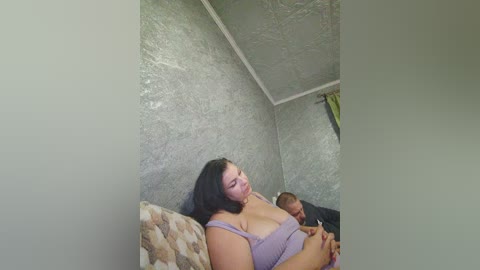 A video of a plus-size woman with shoulder-length black hair, wearing a lavender bra, lying on a beige-patterned pillow in a dimly lit room with textured gray walls and a ceiling fan.