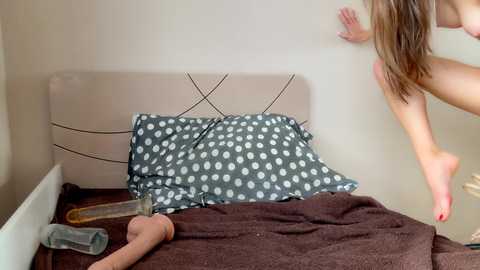 A video of a nude woman with long brown hair, standing on a bed with a beige headboard, holding a large dildo. She is reaching towards a beige wall.