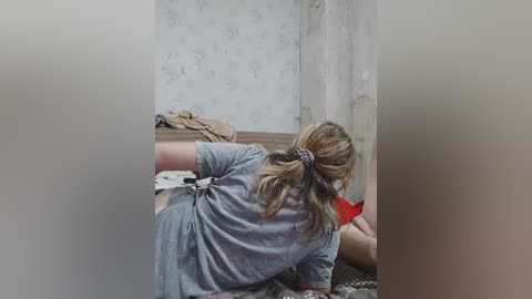 Video of a young woman with long, blonde hair in a ponytail, wearing a grey shirt, kneeling on a bed with floral-patterned wallpaper in the background.