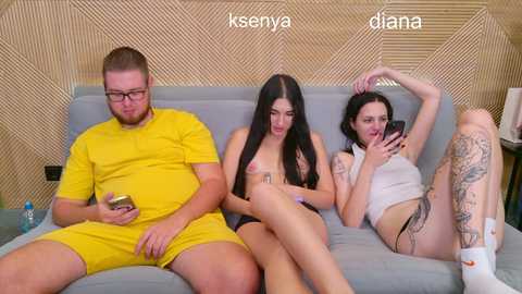 Video: Three young adults, two women and one man, sitting on a light gray couch, each holding a smartphone, in a modern, geometric-patterned room.