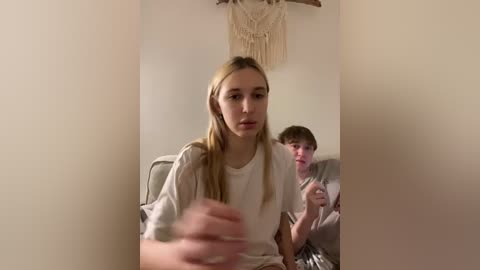 Video of a blonde girl in a white shirt, looking towards a boy in a gray shirt, both seated in a beige room with a macram\u00e9 wall hanging.