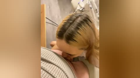 Video of a blonde woman in a beige knit sweater, kneeling, performing oral sex on a man's erect penis. Background shows a light-colored room with a wooden bed frame and a white bedsheet.