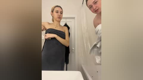Video of a blonde woman in a black towel, standing in a bathroom with a man in a white towel reflected in the mirror.