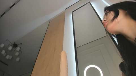 Video of a woman with long black hair, wearing glasses, looking up, in a modern room with light wood paneling, a mirror, and a round light fixture.