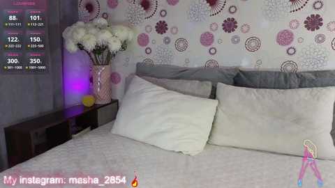 Video of a modern bedroom with a grey bed, white pillows, pink floral wallpaper, a vase with white flowers, and a digital clock displaying 8:01.