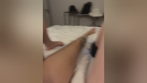 A blurry video shows a person's legs in white socks and a black shoe on a white bed, with a white sheet, in a dimly lit, sparsely furnished room.