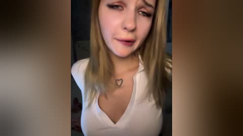 Video of a young blonde woman with a heart tattoo on her neck, wearing a white V-neck shirt, looking serious and pensive.