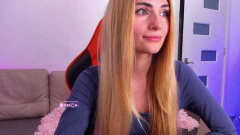 Video of a young woman with long blonde hair, fair skin, and green eyes, wearing a blue top, seated indoors with a beige couch and a red gaming chair in the background.