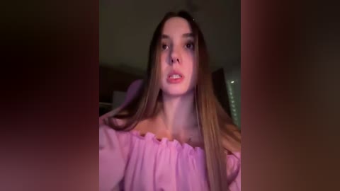 A video of a young Caucasian woman with long, straight brown hair, wearing a light pink off-shoulder top, indoors with dim lighting.