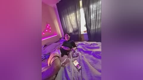 A video of a young person with a medium build, wearing a black shirt and glasses, lying on a bed with a smartphone in hand. The room is dimly lit with purple lights, and there are sheer curtains and a window visible in the background.