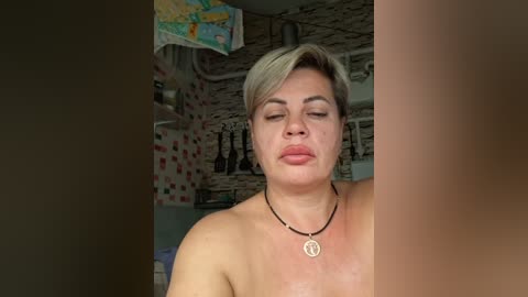 Video of a topless, short-haired woman with fair skin, wearing a black necklace. She has a neutral expression. Background shows a cluttered kitchen with a map, utensils, and colorful wallpaper.