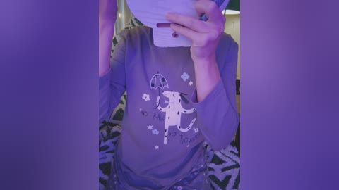 Video of a person in a gray T-shirt with a cartoon elephant design, holding a smartphone, taken indoors with a purple light effect.