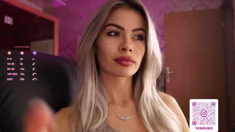 Video of a young woman with platinum blonde hair, fair skin, and a slender build, wearing a light-colored top, captured indoors with a calendar and QR code overlay.