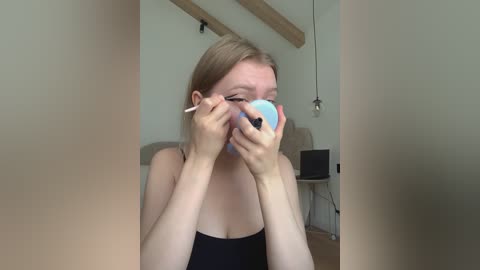 Video of a young blonde woman applying makeup in a minimalist room with light walls, a wooden beam, and hanging lightbulbs. She wears a black tank top, holding a blue eyeshadow palette and brush.