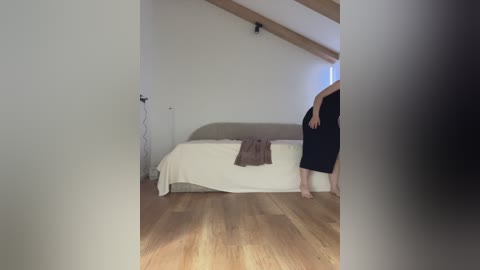 A video captures a person in a black dress, standing near a bed with white sheets and a brown blanket in a dimly lit, minimalist attic bedroom with light wooden floors.