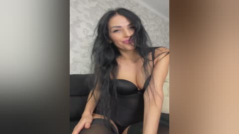 Video of a seductive woman with long black hair, wearing a black, strapless bodysuit, sitting on a couch against a gray and white patterned wall.