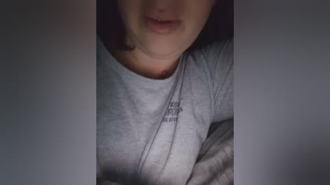 A close-up video of a person's face and chest wearing a gray, long-sleeved shirt with a small, faintly visible logo. The background is dark, with blurry, indistinct shapes. The person has fair skin and a neutral expression.