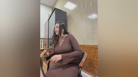 Video of a plus-sized woman with long black hair, wearing a brown, long-sleeved dress with cutouts, seated in a barber chair. The room has beige walls, a wooden panel, and a black screen.