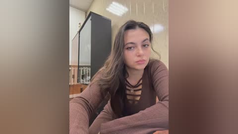 Video of a young woman with long, wavy brown hair, wearing a brown ribbed sweater, leaning on a wooden table in a dimly lit room with cream-colored walls and a radiator.