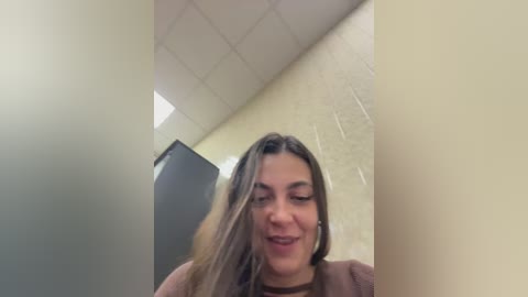 A video of a young woman with long, light brown hair, wearing a brown sweater, smiling slightly, in a dimly lit room with beige walls and a fluorescent ceiling.