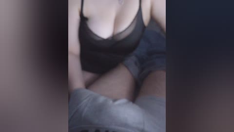 A video shows a close-up of a woman with fair skin, wearing a sheer black lace bra and gray shorts, seated on a bed with dim lighting. The image is slightly blurred.