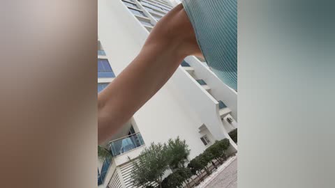 Video of a close-up, high-angle view of a person's arm and hand wearing a turquoise sleeveless shirt, against a modern, white, multi-story building with glass windows and green shrubbery.