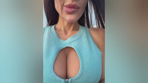Video of a woman with long brown hair, wearing a light blue ribbed crop top with a deep neckline revealing ample cleavage. Her skin is tan, and she has full lips. Background is a blurred gradient of light and dark tones.