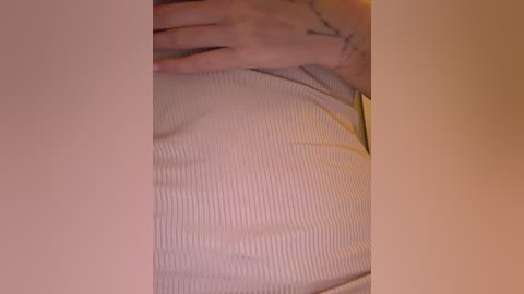 A close-up video of a person's hand resting on a ribbed, light-colored sweater. The sweater has a textured, ribbed pattern, and the person's fingers are visible, showing a tattoo. The background is out of focus, with a warm, ambient light.