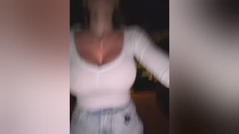 Video of a woman in a white, long-sleeved, form-fitting top, with visible cleavage, and high-waisted, light blue jeans. The background is blurred, making her the focal point.