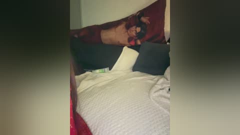Video of a woman with short dark hair lying on a bed with a red and black comforter, wearing a white t-shirt, and partially covered by a white blanket.
