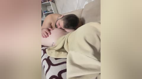 Video of a pregnant woman lying on a beige couch, covered by a beige blanket. She has short dark hair, fair skin, and a small tattoo on her upper arm. Her partner, with a shaved head, is kissing her belly. Background shows a white wall and a shelf with various items.