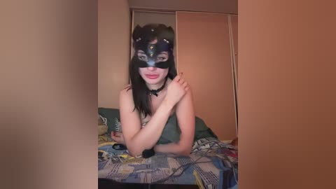 Video of a young woman with fair skin, wearing a black cat mask, black choker, and green top, lying on a colorful quilted bedspread in a dimly lit, beige-walled room with a closed wardrobe in the background.