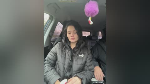 Video of a young woman with long black hair, wearing a puffy gray jacket, sitting in a car with a pink air freshener hanging from the rearview mirror.