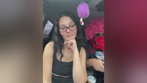 Video of a young woman with long black hair, glasses, and a black tank top, seated in a car, holding a pen, next to a man in a red wig and mask.