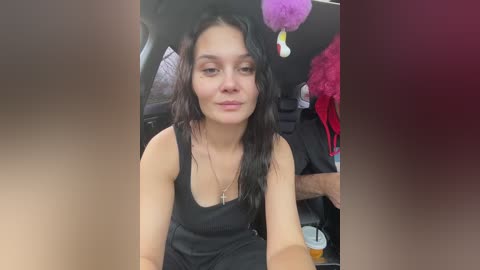 Video of a young woman with long, wavy black hair, wearing a black tank top, sitting in a car, with a purple stuffed animal on the seat behind her.