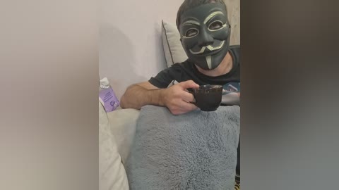 Video of a man in a black mask with a white mustache and goatee, wearing a gray shirt, holding a black coffee mug on a gray blanket, leaning against a white wall.