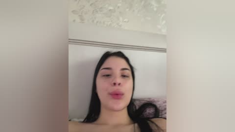 A video of a young woman with light skin and long black hair, making a pouty face, standing in a room with a floral-patterned ceiling and a bed with a pink blanket.