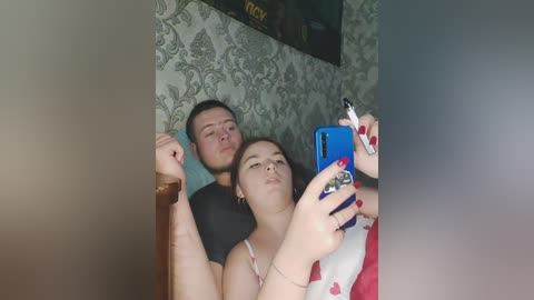 Video of a young couple in a dimly lit room, with a man resting his head on a woman's shoulder, both holding a phone for a selfie.