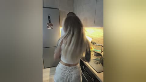 A video of a woman with long, straight, blonde hair, wearing a white crop top and white shorts, cooking in a modern kitchen with stainless steel appliances and wooden countertops.