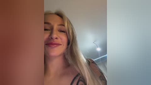 Video of a young, fair-skinned blonde woman with long hair, wearing a black top, smiling slightly. Background shows a dimly lit room with a ceiling light and exposed beams.