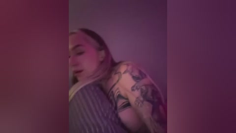 Video of a light-skinned woman with long, blonde hair, wearing a striped top and revealing black bra, with a prominent tattoo sleeve. The image is dimly lit with a purple hue, giving a moody, intimate atmosphere.