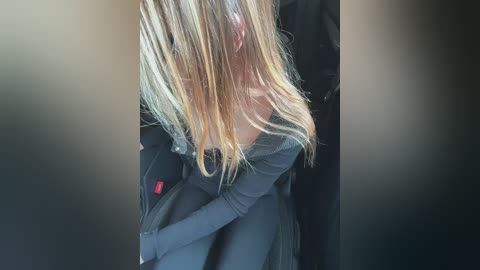 Video of a young woman with long blonde hair, wearing a black off-shoulder top, hugging herself in a dimly lit room.