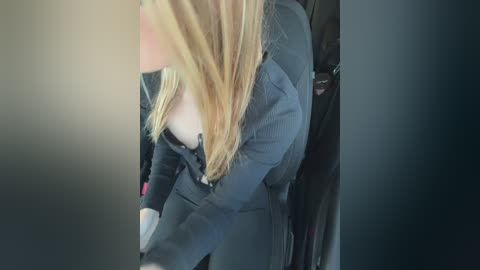 A video captures a woman with long blonde hair, wearing a dark blazer, adjusting her bra inside a car. Her face is blurred, focusing on her chest and arms.